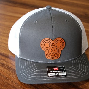 May include: Gray and white trucker hat with a brown leather patch featuring a Celtic knot design. The hat has a Richardson 112 adjustable strap.