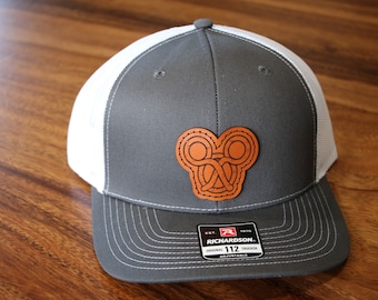 May include: Gray and white trucker hat with a brown leather patch featuring a Celtic knot design. The hat has a Richardson 112 adjustable strap.
