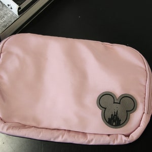 K&ouml;nnte beinhalten: Eine rosa Bauchtasche mit einer schwarzen Mickey-Maus-Silhouette, die das Disney-Schloss zeigt. Die Tasche hat einen Rei&szlig;verschluss und einen verstellbaren Gurt.