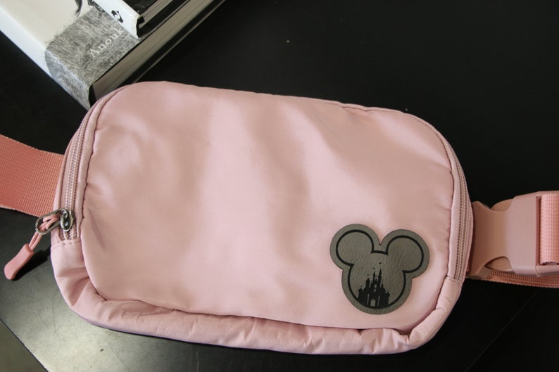 K&ouml;nnte beinhalten: Eine rosa Bauchtasche mit einer schwarzen Mickey-Maus-Silhouette, die das Disney-Schloss zeigt. Die Tasche hat einen Rei&szlig;verschluss und einen verstellbaren Gurt.