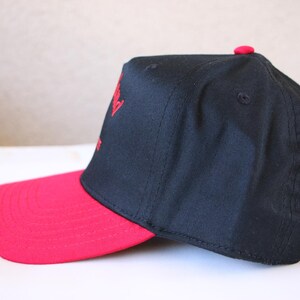 K&ouml;nnte beinhalten: Eine schwarze und rote Baseballkappe mit rotem Schirm und einem roten gestickten Logo auf der Vorderseite.