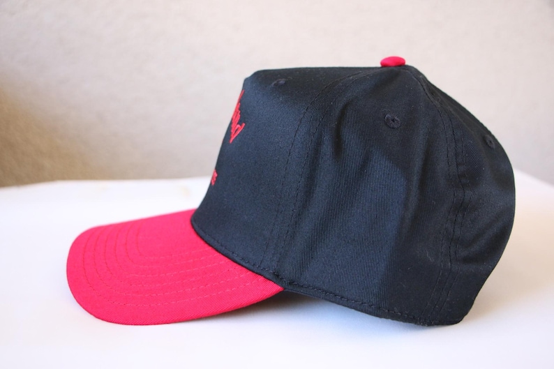 K&ouml;nnte beinhalten: Eine schwarze und rote Baseballkappe mit rotem Schirm und einem roten gestickten Logo auf der Vorderseite.
