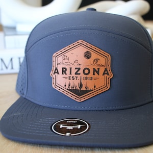 Puede incluir: Una gorra de béisbol azul oscuro con un parche de cuero marrón que dice "Arizona - Est. 1912" con un gráfico de un paisaje desértico.