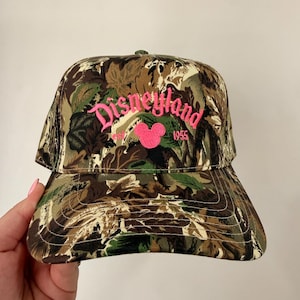 Peut inclure: Casquette de baseball camouflage avec le mot "Disneyland" en rose, une tête de Mickey Mouse rose et l'année "1955". La casquette présente un motif de feuilles marron, vert et beige.