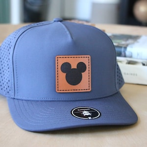 May include: A blue baseball cap with a perforated side panel and a curved brim. The cap features a square, brown leather patch with a black Mickey Mouse silhouette. A black and white sticker is on the brim.