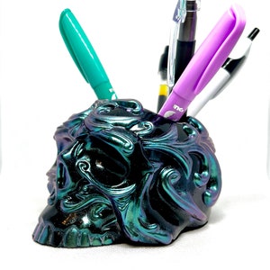 May include: A black skull-shaped desk organiser with iridescent green and purple paint. The skull has ornate details and is holding four pens, including a purple pen with the word "Ink" on it.