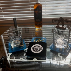 May include: Three clear glass tumblers with etched designs on a glass table. The tumblers are on blue coasters with gold bullet casings. A bottle of Fiddler Bourbon Whiskey is in the center of the table. A small wooden model of a rifle is on a tumbler to the right.