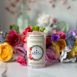 May include: A cylindrical deodorant stick with a clear cap, labeled "Strawberry Marshmallow Cream Baking Soda Free Deodorant." The label features strawberries and a clock design. The product is surrounded by colorful flowers.