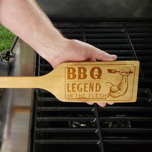 May include: A bamboo BBQ grill scraper with the text "BBQ LEGEND IN THE FLESH" and an illustration of a muscular arm holding a fork. The scraper is held over a grill.