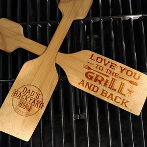 May include: Two wooden grilling spatulas with engraved text. One spatula reads "Dad's Backyard BBQ" and the other says "Love You to the Grill and Back." The spatulas are light brown and rest on a black grill.