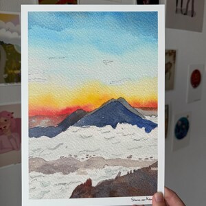 Può includere: Dipinto ad acquerello di una catena montuosa al tramonto. Il cielo passa dal blu al giallo e al rosso, con montagne blu scuro e nuvole bianche. La firma dell'artista, Sharon van Kouwen, è in basso a destra.
