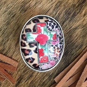 May include: A round, silver-toned, decorative pin with a cross design. The cross is red and white with a floral pattern and a leopard print background.
