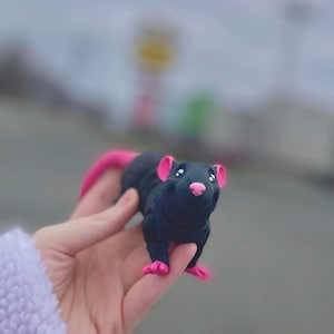 Pode incluir: Estatueta de rato preto feita à mão com orelhas, nariz, pés e cauda rosa. O rato tem olhos brancos pequenos e é segurado em uma mão contra um fundo desfocado ao ar livre. A estatueta parece ser feita de argila ou material semelhante.