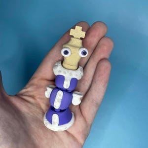 Op de afbeelding: Een grillig 3D-geprint schaakkoning-beeldje. De koning heeft een lichtbruin hoofd met wiebelogen, een witte kraag en een paars lichaam met witte accenten. Het beeldje wordt in een hand gehouden tegen een blauwe achtergrond.