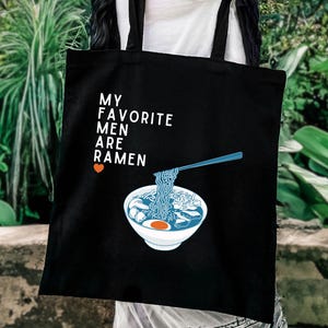May include: A black tote bag featuring a white graphic of a bowl of ramen with blue noodles being lifted by chopsticks. The text reads MY FAVORITE MEN ARE RAMEN with a small orange heart icon. The bag has two black shoulder straps for carrying.