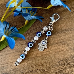 May include: A silver-toned keychain with a beaded charm featuring blue evil eye beads, white beads, and silver flower accents. A silver Hamsa hand charm hangs from the end. Blue artificial flowers are in the background.