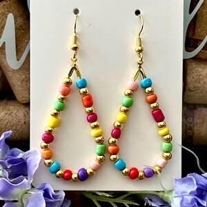 May include: A pair of teardrop-shaped earrings featuring a gold-toned wire frame strung with alternating small metallic gold beads and multi-colored opaque beads in shades of pink, blue, yellow, green, orange, red, and purple.