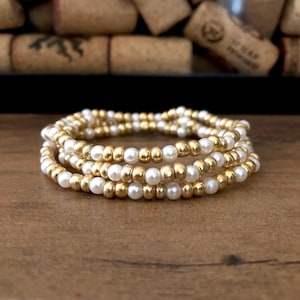 May include: A stack of four beaded bracelets. Each bracelet alternates between small, gold-colored beads and white, pearl-like beads. The bracelets are displayed on a wooden surface, with wine corks in the background.
