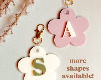 May include: Two flower-shaped keychains with gold clasps. The keychain on the left is white with a green letter "S" in the center. The keychain on the right is pink with a white letter "A" in the center. The text "more shapes available!" is written below the keychains.