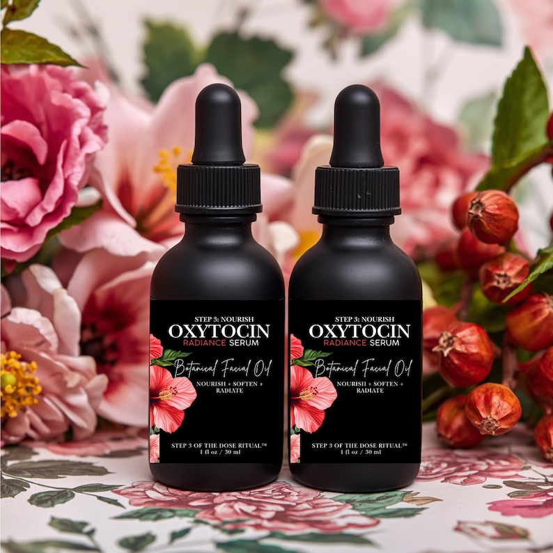 May include: Two black bottles of Oxytocin Radiance Serum with a botanical facial oil label. The label includes a red hibiscus flower illustration. The bottles are 1 fl oz / 30 ml each. The background features pink flowers and foliage.
