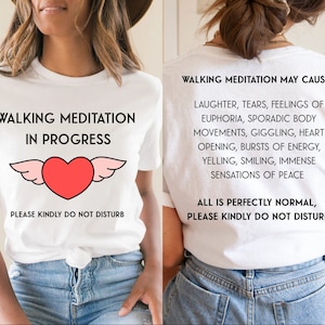 Può includere: T-shirt bianca con la scritta "WALKING MEDITATION IN PROGRESS" e un cuore con le ali. Il retro della maglietta elenca i possibili effetti della meditazione camminata. Il testo "PLEASE KINDLY DO NOT DISTURB" appare su entrambi i lati.