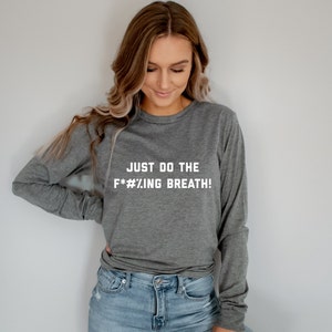 May include: A heather gray long-sleeve shirt with the text "JUST DO THE F*#%.ING BREATH!" in white block letters. The shirt is made of a soft material and has a crew neck.