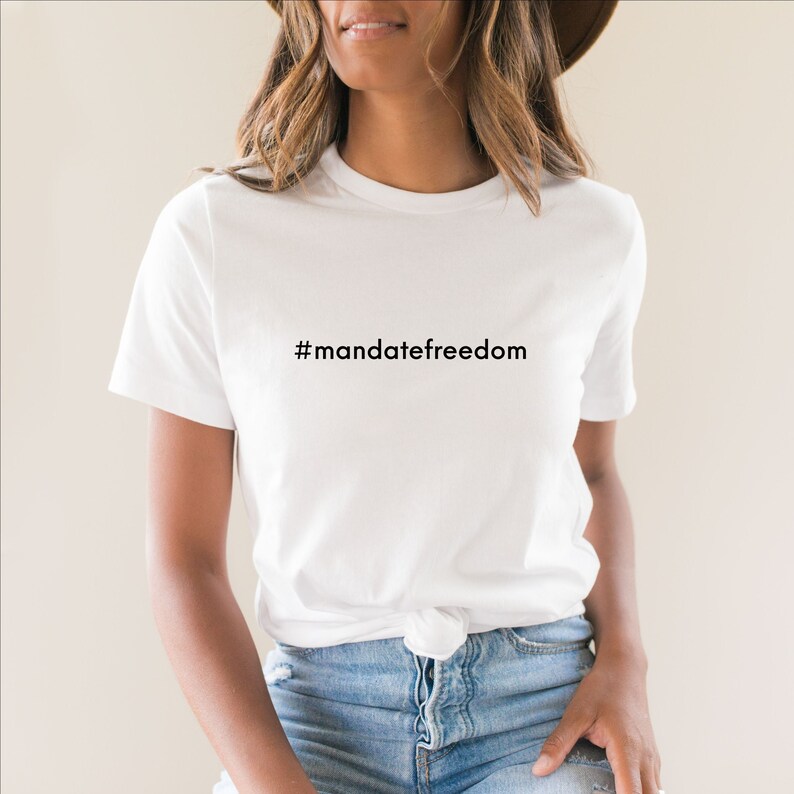 May include: White t-shirt with the text "#mandatefreedom" in black. The shirt is a classic crew neck style and is worn with blue jeans. The overall look is casual and comfortable.