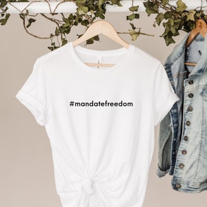 May include: A white t-shirt with the text "#mandatefreedom" in black. The shirt is hanging on a wooden hanger. A denim jacket hangs to the right. Green vines are draped above the shirt.