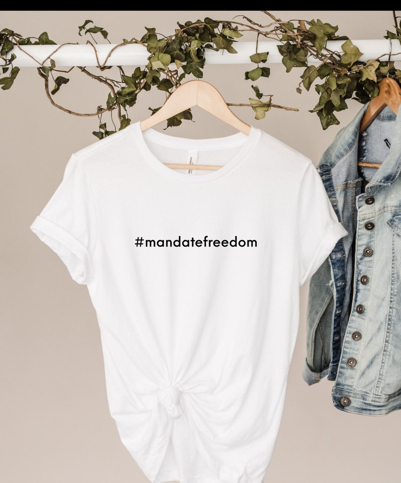 May include: A white t-shirt with the text "#mandatefreedom" in black. The shirt is hanging on a wooden hanger. A denim jacket hangs to the right. Green vines are draped above the shirt.