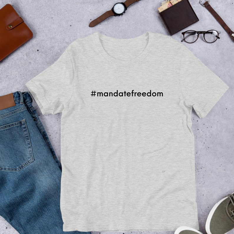 May include: Light gray t-shirt with the text "#mandatefreedom" in black. The shirt is made of a soft material and is displayed on a flat surface with other items, including a watch, wallet, and jeans.