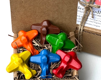 Puede incluir: Una caja de cartón llena de seis pequeños crayones con forma de avión en varios colores, incluyendo rojo, azul, amarillo, verde, naranja y marrón. La caja está atada con una cuerda y tiene una etiqueta que dice "Bee Doodle Coloring Crayons".