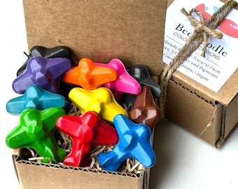 Puede incluir: Una caja llena de lápices de colores con forma de avión de colores. Los lápices de colores están hechos con cera de granja natural y pigmentos. La caja es de cartón marrón y tiene una etiqueta que dice "Beepodle Crayons".