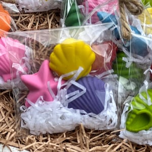 Könnte beinhalten: Eine Sammlung bunter Seifen in Muschelform in transparenten Zellophanbeuteln, die mit Bindfäden gebunden sind. Die Seifen sind in verschiedenen Farben erhältlich, darunter Rosa, Gelb, Lila und Grün. Die Seifen sind in weißen Papierschnipseln eingebettet.