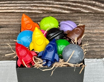 May include: A black box filled with colorful crayons shaped like rockets and planets. The crayons are arranged in a decorative way and are ready for creative play.
