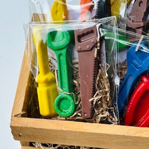 May include: A wooden crate filled with brown packing paper and various colourful toy tools wrapped in clear plastic bags. The tools include a wrench, a saw, a screwdriver, and pliers.