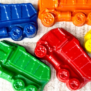 May include: Five toy dump trucks made of wax crayons. The trucks are blue, green, red, orange, and yellow. Each truck has a detailed design with a bed, wheels, and a cab.