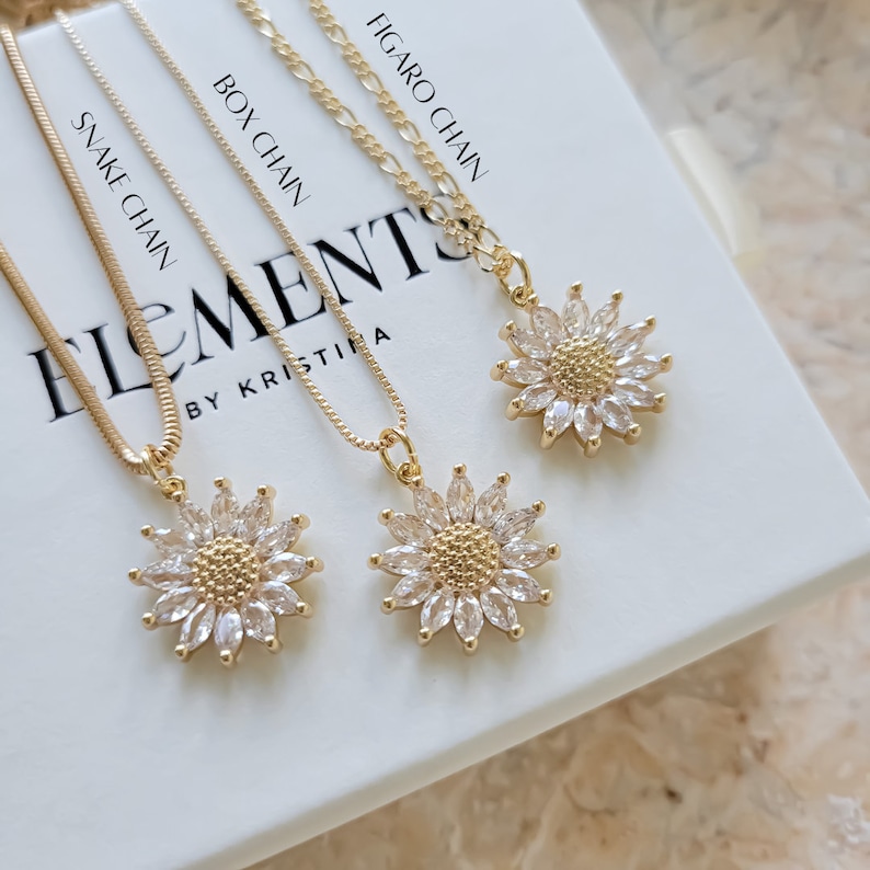 May include: Three gold necklaces with different chain styles: snake, box, and figaro. Each necklace features a small sunflower pendant with clear cubic zirconia stones.