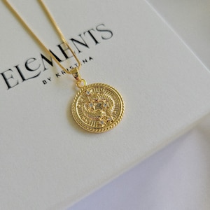Puede incluir: Collar de oro con un colgante redondo que presenta un dise&ntilde;o detallado y peque&ntilde;as piedras transparentes. El colgante est&aacute; sobre un fondo blanco con el texto "Elements by Kristina".