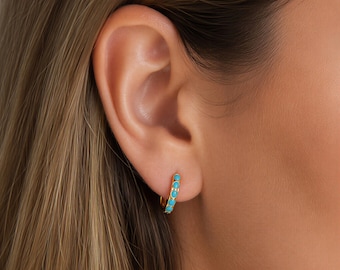 May include: A small gold hoop earring featuring a row of five round, bright turquoise stones set along the outer edge. The earring is secured through the earlobe, showcasing a polished metallic finish and vibrant blue-green circular accents.