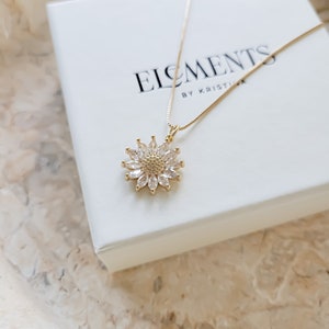 May include: A gold necklace with a clear crystal sunflower pendant. The necklace is on a white box with the text "Elements by Kristia" printed on it.