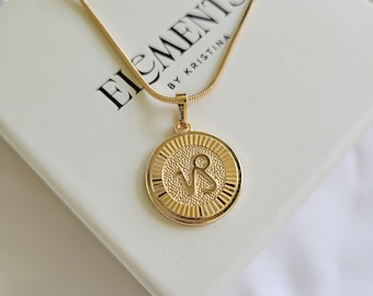 May include: Gold necklace with a round pendant featuring the Capricorn zodiac symbol. The pendant has a textured surface and a raised border. The necklace is on a white box with the text "Elements by Kristina".