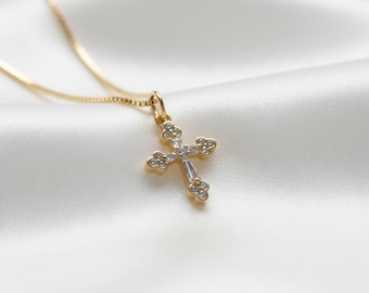 May include: A gold-colored necklace with a cross pendant. The cross is adorned with clear, sparkling gemstones. The necklace is displayed on a white, silky fabric, creating a luxurious and elegant presentation.