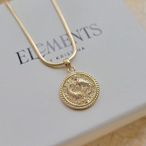 Puede incluir: Collar de cadena dorada con un colgante redondo que presenta un s&iacute;mbolo zodiacal de dos peces con peque&ntilde;as piedras transparentes. El colgante est&aacute; sobre una caja blanca con el texto "ELEMENTS BY KRISTINA".