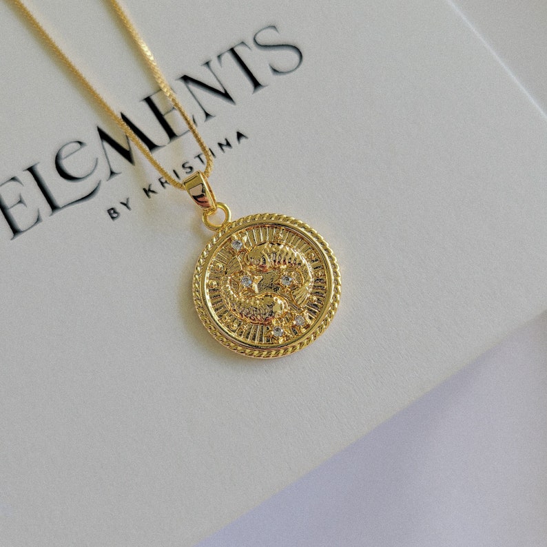 Puede incluir: Collar de oro con un colgante redondo que presenta un dise&ntilde;o detallado de dos peces y peque&ntilde;as piedras transparentes. El colgante est&aacute; en una cadena con un cierre de langosta. El collar se muestra sobre un fondo blanco con el texto "ELEMENTS BY KRISTINA".