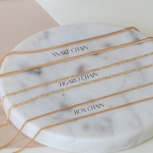 Puede incluir: Tres cadenas de oro sobre una superficie de m&aacute;rmol blanco. Las cadenas est&aacute;n etiquetadas como "Snake Chain", "Figaro Chain" y "Box Chain".