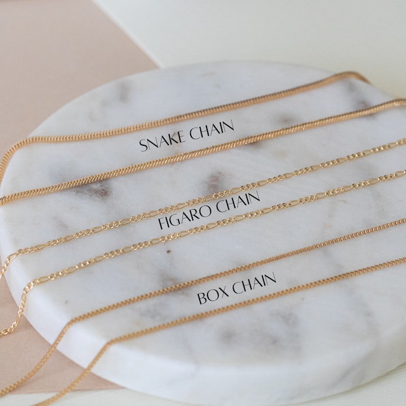 Puede incluir: Tres cadenas de oro sobre una superficie de m&aacute;rmol blanco. Las cadenas est&aacute;n etiquetadas como "Snake Chain", "Figaro Chain" y "Box Chain".