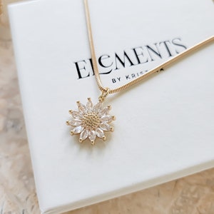May include: A gold chain necklace with a clear crystal flower pendant. The pendant is a daisy with a gold center and clear crystal petals. The necklace is on a white box with the text "ELEMENTS BY KRISTA" printed on it.