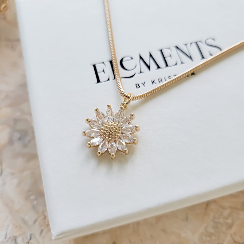 May include: A gold chain necklace with a clear crystal flower pendant. The pendant is made of small, faceted crystals that are set in a gold metal setting. The necklace is on a white background with the text "ELEMENTS BY KRISTA" printed on it.