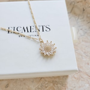 May include: A gold chain necklace with a sunflower pendant featuring clear cubic zirconia stones. The necklace is displayed on a white box with the text "Elements by Kristina".