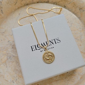 Puede incluir: Un collar de cadena dorada con un colgante redondo que presenta un s&iacute;mbolo del zod&iacute;aco y peque&ntilde;as piedras brillantes. El collar se muestra en una caja blanca con el texto "ELEMENTS BY KRISTINA".
